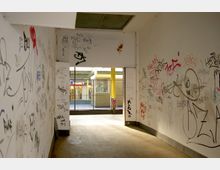 Ein verlassener Gang mit weiß gestrichenen Wänden, die mit verschiedenen Graffiti und Kritzeleien bedeckt sind. Am Ende des Ganges ist ein Gittertor sichtbar, das den Blick auf eine Einkaufspassage mit einem Markt-Schild freigibt.