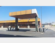 Außenansicht einer Raststätte mit orange-brauner Fassade und kantiger Architektur, einschließlich Überdachung auf Pfeilern. Im Hintergrund befinden sich ein klarer, blauer Himmel sowie weitere Gebäude und Freiflächen.