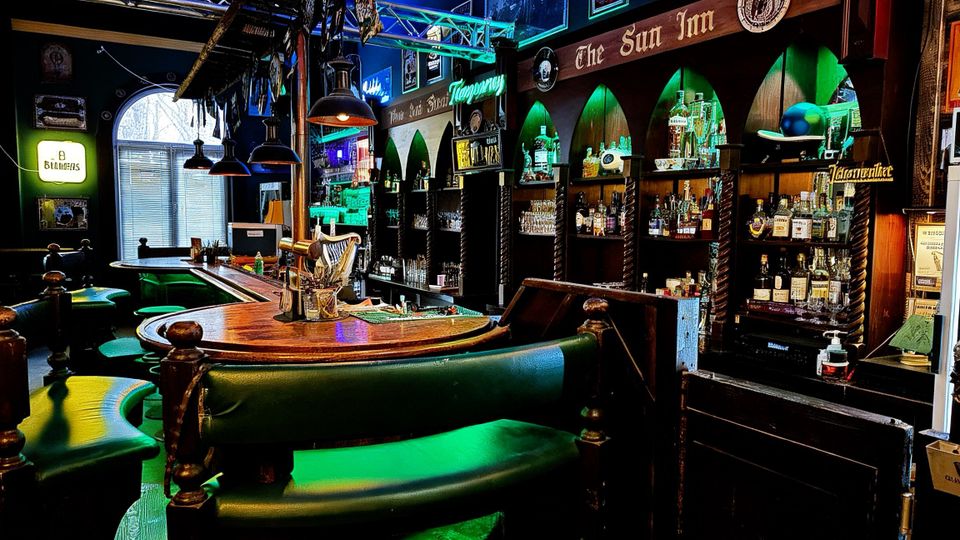 Innenraum einer Bar mit gebogener Holztresenanlage, grünen Polsterbänken und Barhockern sowie hängenden Industrielampen und Metallträgern an der Decke. An der Rückwand stehen beleuchtete Regale mit Flaschen und Gläsern in bogenförmigen Nischen, umgeben von Schildern und Neonwerbung.