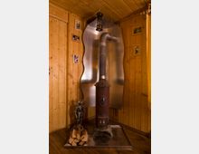 Ecke eines rustikalen Raumes mit Holzvertäfelung, einer roten, verzierten Gusseisen-Kaminofen mit Metallrohr, das in die Wand führt, sowie einem Holzstapel und dekorativer Ritterfigur auf einem Metallsockel. Im Hintergrund hängen kleine gerahmte Bilder an der Wand.