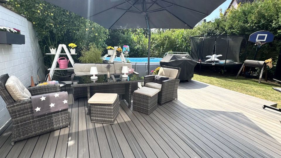 Große Holzterrasse im Garten mit Rattan-Loungegruppe und Esstisch unter einem breiten Sonnenschirm. Im Hintergrund stehen ein aufgestellter Pool, ein abgedeckter Grill sowie ein Trampolin und ein Basketballkorb vor hohen Hecken.