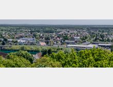 Weitläufige Ansicht einer Kleinstadt mit Wohnhäusern und Industriegebäuden, umgeben von grünen Bäumen und Wäldern am Horizont. Im Vordergrund dichte Baumkronen, dahinter eine Straße und verschiedene Gebäude mit roten und grauen Dächern.