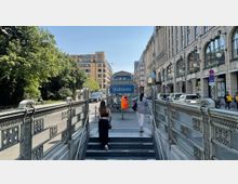 Treppenaufgang einer U-Bahn-Station mit der Beschilderung „Stadtmitte“, flankiert von historischen Geländern. Im Hintergrund sind ein modernes Bürogebäude, eine Baumreihe sowie eine belebte Straße mit Autos und Fußgängern zu sehen.