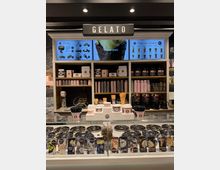 Eine Eisdiele mit einer Glasvitrine, die verschiedene Eissorten präsentiert. Im Hintergrund befinden sich Regale mit Eiswaffeln, Pappbechern und Dekoration sowie digitale Bildschirme, die Eissorten und Getränke bewerben.