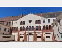 Historisches Gebäude mit roten Backsteinakzenten und gotisch geformten Fenstern in einem hell verputzten Bereich. Im oberen Teil der Fassade befindet sich ein Wappen, umgeben von einem roten Ziegeldach und mehreren Fenstern.