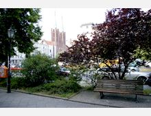 Eine städtische Szene mit einer Parkbank im Vordergrund, umgeben von Büschen und Bäumen. Im Hintergrund sind eine belebte Straße, historische Gebäude und eine große Backsteinkirche mit hohen Türmen erkennbar.