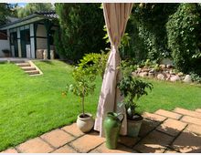 Ein gepflegter Garten mit grünem Rasen, einem kleinen weißen Gebäude mit Fachwerkelementen und Treppenstufen im Hintergrund. Im Vordergrund stehen drei Pflanzen in Töpfen und ein grün glasierter Krug auf einer Terrasse mit beigefarbenen Steinplatten.