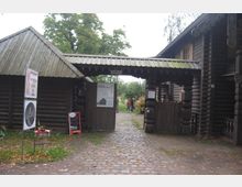 Holzgebäude mit traditioneller russischer Bauweise und überdachtem Eingangstor, das zu einem Innenhof führt. Links stehen Schilder für ein Museum und Café, umgeben von Kopfsteinpflaster und grüner Vegetation.