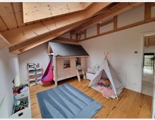 Ein Kinderzimmer unter einem Dach mit sichtbaren Holzbalken. Es enthält ein Spielhaus aus Holz, ein weißes Spielzelt, eine Hängeschaukel und Regale mit Spielzeug und Büchern, auf einem Holzboden mit einem blauen Teppich.