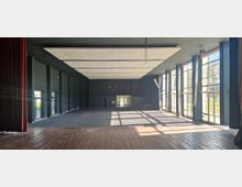 Ein leerer Veranstaltungsraum mit einer erhöhten Bühne aus Holz im Vordergrund, hohen Fenstern an der rechten Seite, die viel Tageslicht hereinlassen, sowie einer dunklen Wand am Ende des Raumes mit einer Doppeltür in der Mitte. Die Decke ist mit rechteckigen Akustikplatten verkleidet.