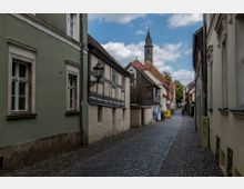 Schmale, gepflasterte Gasse in einer historischen Altstadt mit Fachwerkhäusern und verputzten Fassaden. Im Hintergrund ist ein Kirchturm mit spitzer Haube zu sehen, umgeben von Bäumen und weiteren Gebäuden.