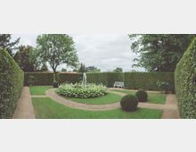 Geometrisch gestalteter Garten mit geschnittenen Hecken, einer kreisförmigen Blumenbeetanlage mit Springbrunnen in der Mitte und einem weißen Holzbänkchen im Hintergrund. Die Wege aus Kies umranden die Beete und führen durch die Grünflächen.