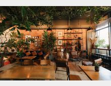 Gemütlicher Innenraum eines Cafés mit Holzmöbeln, einer Ziegelwand, Pflanzen und Dekorationen im Vintage-Stil. Große Fenster auf der rechten Seite lassen Tageslicht herein und verleihen dem Raum eine helle Atmosphäre.