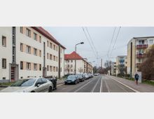 Straßenszene mit mehrstöckigen Wohngebäuden im Hintergrund, teils mit roten Dächern und Balkonen; parkende Autos entlang der Straße und daneben verlaufende Straßenbahngleise. Ein Gehweg und Stromleitungen für die Straßenbahn sind ebenfalls zu sehen.