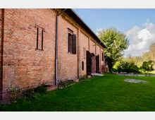 Backsteinbau mit großen Holztüren und einfachen Backsteinverzierungen, umgeben von einer grünen Wiese und Bäumen im Hintergrund. Neben dem Gebäude befinden sich Sitzgelegenheiten und ein gepflasterter Weg.