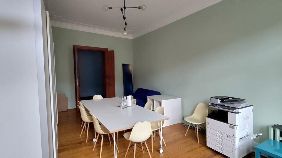 Helles Besprechungszimmer mit langen weißen Tisch und mehreren Stühlen auf Holzboden, mintgrünen Wänden und hoher Decke mit moderner Deckenleuchte. Rechts steht ein großer Kopierer neben einem Heizkörper, hinten eine Holztür und einige Büromöbel.