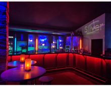 Innenraum einer modernen Bar mit U-förmigem Tresen, rot hinterleuchteter Front und beleuchteten Regalen voller Flaschen und Gläser. Neonfarbene Lichtakzente in Blau, Grün und Orange sowie kleine Tische mit Kerzenlicht prägen die Lounge-Atmosphäre.