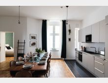 Helle, moderne Wohnküche mit Esstisch, der für mehrere Personen gedeckt ist, grauen Küchenschränken und einer Arbeitsfläche. Große Fenster mit dunklen Vorhängen lassen Tageslicht herein, und der Raum hat Holzboden sowie Fliesen im Küchenbereich. Ein angrenzendes Schlafzimmer ist durch eine offene Tür sichtbar.