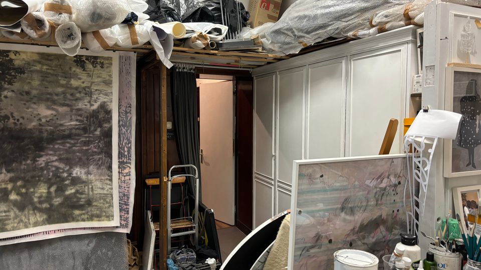 Vollgestellter Atelier- bzw. Lagerraum mit großen gerollten Leinwänden und Verpackungsmaterial auf hohen Regalen, einem großen Bild an der linken Wand und weißen Einbauschränken rechts. Im Vordergrund stehen Arbeitsutensilien wie Farben, Pinsel und Behälter auf einem Tisch; hinten führt eine offene Tür in einen schmalen Nebenraum.
