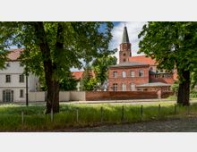 Backstein-Gebäude mit einem spitzen Kirchturm im Hintergrund, umgeben von Bäumen und einer niedrigen Ziegelmauer. Im Vordergrund befindet sich eine gepflasterte Straße mit einer schmalen Grünfläche und einem Zaun.