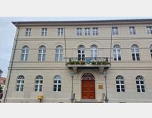 Historisches Gebäude mit hellbeiger Fassade, symmetrischen Rundbogenfenstern und einem hölzernen Eingangstor. Über dem Eingang befindet sich ein kleiner Balkon mit Blumen und einem Wappen, flankiert von zwei Laternen.
