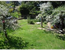 Grüne Gartenfläche mit blühenden Obstbäumen und Streuwiese, umgeben von einem Holzzaun; im Hintergrund dichter Baumbestand. Im Vordergrund befinden sich ein Sandkasten und ein kleiner Spielbereich mit Spielzeug.