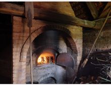 Traditioneller Backofen aus Ziegelsteinen in einer rustikalen Umgebung, mit einem brennenden Feuer im Inneren und Holzstapeln im Vordergrund. Die Konstruktion umfasst einen gewölbten Eingang und Holzbalken unter einem offenen Dach.