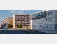 Mehrgeschossige Wohngebäude mit Balkonen und schlichter Architektur stehen an einer ruhigen Straße; daneben ein Gebäude mit grauer Fassade, großen Fensterflächen und Graffiti an der Wand. Einige Bäume und Grünflächen trennen die Bauwerke.