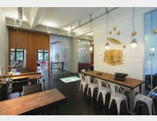 Arbeitsbereich in modernem Loft-Stil mit Holztischen, schwarzen und weißen Stühlen sowie Hängelampen. Im Hintergrund sichtbar: Räume mit Glaswänden, ein Wandbild und ein Holzboden.