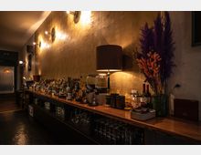 Ein stilvoll eingerichteter Bartresen mit einer goldfarbenen Wand im Hintergrund, beleuchtet von warmen Wandlampen. Auf der Theke stehen diverse Spirituosenflaschen, Gläser, Dekorationsmaterialien und eine Tischlampe, neben einem Trockenblumenarrangement in kräftigen Farben.