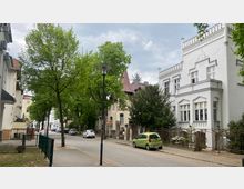 Eine ruhige Straße in einem Wohngebiet mit Bäumen gesäumt, links und rechts stehen historische Gebäude mit verzierten Fassaden. Im Vordergrund ist ein weißes Haus mit der Jahreszahl "1905" über dem Eingang zu sehen; geparkte Autos stehen entlang der Straße.