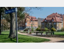 Ein kleiner Park mit Grünflächen, einem geschwungenen Weg, Parkbänken und einem Wegweiser mit der Aufschrift „Karl-Marx-Platz“. Im Hintergrund stehen mehrstöckige Wohnhäuser mit roten Ziegeldächern und hellen Fassaden, umgeben von Bäumen und Sträuchern.