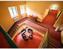 Treppenhaus in einem Gebäude mit gelben Wänden und roten Teppichböden, zentraler Sitzgruppe mit Sesseln und kleinem Tisch im unteren Bereich, grün-geländerter Treppe und sichtbaren Heizkörpern unter Fenstern. Ein Flur mit Notausgangsschild führt von der oberen Ebene weg.