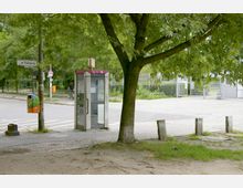 Öffentlicher Platz mit einer alten Telefonzelle, umgeben von Gehwegen, Bäumen und Grünflächen. Im Hintergrund sind eine Straße mit Markierungen, ein Metallzaun und Menschen auf Fahrrädern erkennbar.