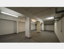 Ein leerer Kellerraum mit weißen Backsteinwänden, mehreren Betonpfeilern und einer niedrigen Decke mit sichtbaren Rohren und Leuchtstofflampen. Der Boden ist glatt und grau, und die Umgebung wirkt industriell.