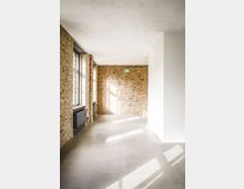 Heller, leerer Innenraum im Loft-Stil mit freiliegenden Backsteinwänden, glatter Betonboden und großer Fensterfront links. Sonnenlicht fällt in den Raum; an der Rückwand ist ein grünes Notausgangsschild sichtbar.