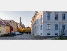 Straßenszene mit gepflasterter Fahrbahn, flankiert von Wohngebäuden mit hellen Fassaden, einem Straßenschild mit der Aufschrift „Fischerstraße“ und geparkten Autos; im Hintergrund ragt der Turm einer Backsteinkirche über gelb gefärbte Herbstbäume.