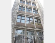 Fassade eines mehrstöckigen Gebäudes mit großen Fenstern in Holzrahmen und einer rustikalen Steinwand auf der linken Seite. Die unteren Fenster befinden sich hinter einem Metallgeländer, und die oberen Stockwerke zeigen eine gleichmäßige Anordnung von Fenstern und Steinverkleidung.