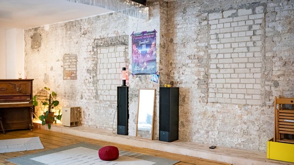Innenraum mit rohen Backstein- und Putzwänden, in denen mehrere zugemauerte Fensteröffnungen sichtbar sind, sowie Holzboden und Deckenbeleuchtung. An der Wand stehen zwei hohe schwarze Lautsprecher und ein Spiegel; links sind ein Klavier und Pflanzen zu sehen.