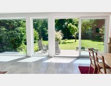Heller Wohn- und Essbereich mit großflächigen bodentiefen Fenstern und einer Schiebetür, die auf eine geflieste Terrasse führen. Draußen ist ein großer, grüner Garten mit Rasen, Bäumen und Sträuchern zu sehen.