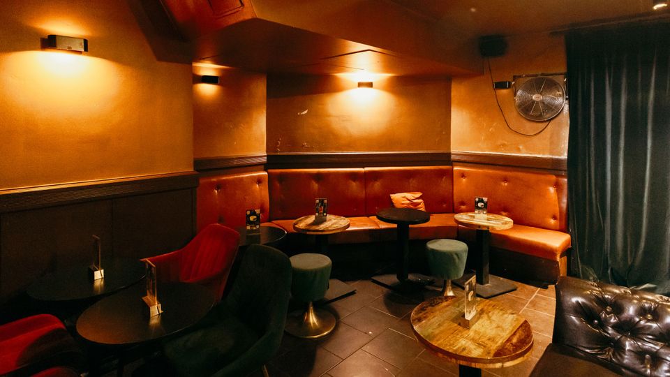 Gemütlicher Bar- oder Lounge-Innenraum mit gepolsterter Eckbank in Braun, kleinen runden Tischen und Hockern auf dunklem Fliesenboden. Warme Wandlampen beleuchten orangefarbene Wände; rechts hängen ein Ventilator und ein schwerer Vorhang.