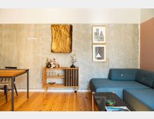 Wohnzimmer mit modernem, reduziertem Design; eine graue Wand mit zwei gerahmten Grafiken und einer goldfarbenen Kunstinstallation. Eine blaue Eckcouch, ein Holztisch mit schwarzem Metallrahmen sowie ein Mid-Century-Servierschrank mit Dekorationen prägen den Raum, der einen Holzboden hat.