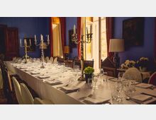 Ein festlich gedeckter langer Esstisch in einem elegant gestalteten Raum mit blauen Wänden, großen Fenstern mit roten Vorhängen und silbernen Kerzenleuchtern als Dekoration. Stühle mit weißen Bezügen umgeben den Tisch, der mit Gläsern, Besteck und Servietten ausgestattet ist.