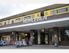 Außenansicht des U-Bahnhofs Ruhleben in Berlin mit gelben U-Bahn-Wagen auf der oberen Ebene, einem großen Schriftzug "RUHLEBEN" an der Fassade und verschiedenen Geschäften sowie einer Uhr im Eingangsbereich darunter. Die Architektur besteht aus Klinkerwänden und einer klar strukturierten Überdachung.