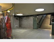 Unterführung mit grauem Boden, grünen Wandfliesen und Treppenaufgang auf der rechten Seite. Links im Bild ist ein Schaufenster mit einer Schaufensterpuppe in rotem Kleid, während die Wände des Durchgangs mit geschlossenen Metallrollläden und Graffiti bedeckt sind.