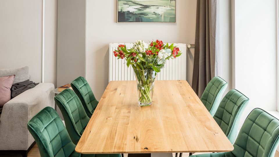 Moderner Essbereich mit langem Holztisch und sechs grünen, gepolsterten Stühlen auf hellem Holzboden. An der Wand hängen ein Landschaftsbild über einem Heizkörper; daneben ein großes Fenster mit dunklen Vorhängen und eine schlichte Pendelleuchte über dem Tisch.