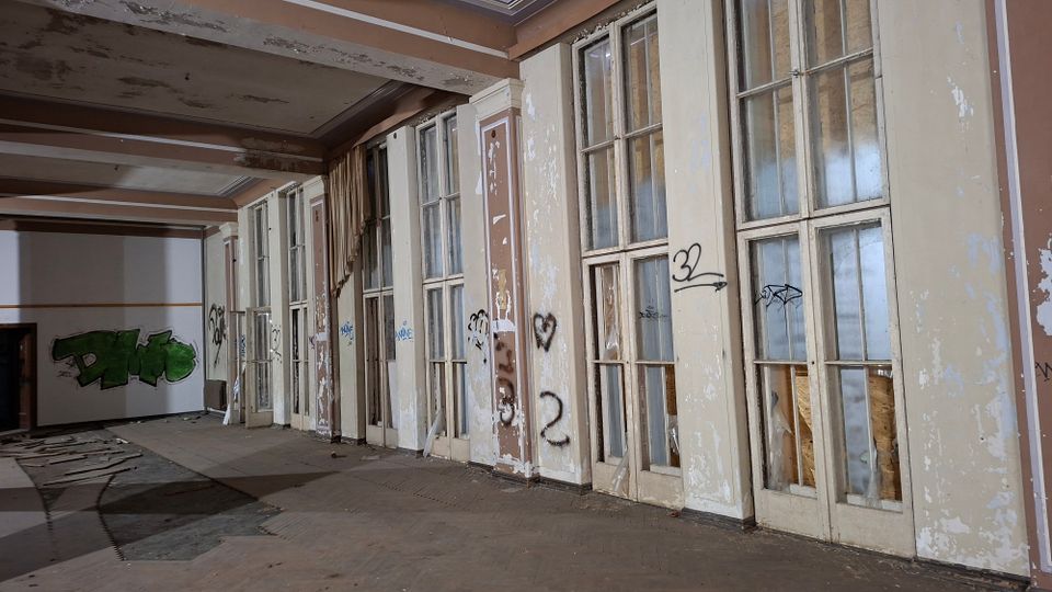 Verlassener Innenraum eines Gebäudes mit einer langen Reihe hoher, mehrfach verglaster Türen und Fenster, teils beschädigt und mit abblätternder Farbe. An Wänden und Türen sind Graffiti zu sehen; der Boden ist stellenweise verschmutzt und mit Trümmern bedeckt.