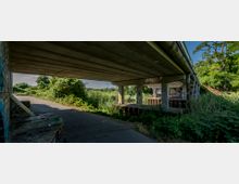 Ansicht einer Brücke aus Beton, die über eine mit Pflanzen bewachsene Fläche und einen asphaltierten Weg führt. Die Brückenpfeiler sind mit Graffiti verziert, und die Umgebung ist von dichtem Grün und Bäumen geprägt.