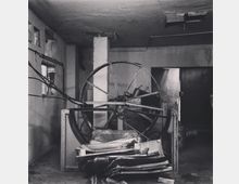 Schwarz-weiß-Aufnahme eines verlassenen industriellen Raums mit einer großen Metallspule im Zentrum, umgeben von Kabeln und gestapeltem Schrott. Die Wände wirken verfallen, mit einem offenen Raum und einer Tür im Hintergrund.