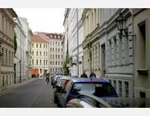 Schmale städtische Straße mit parkenden Autos auf beiden Seiten, gesäumt von historischen Gebäuden mit verzierten Fassaden. Im Hintergrund sind mehrstöckige Wohnhäuser zu sehen, Passanten und ein Fahrradfahrer beleben die Szene.